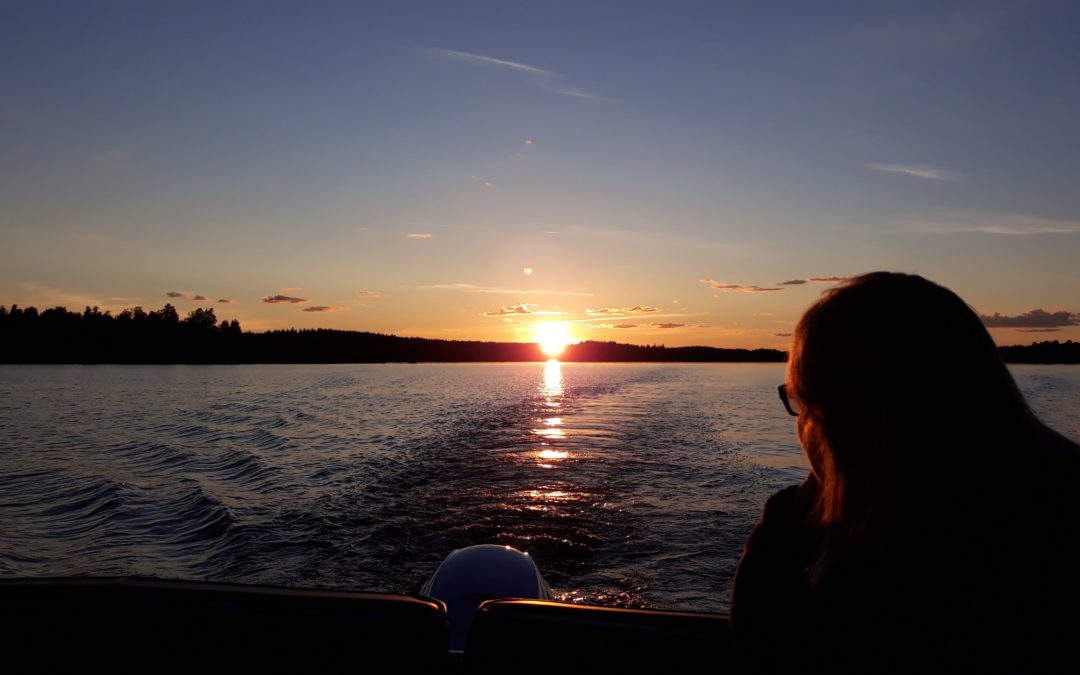 Mari Lång-Kauppi järvellä veneessä katselee auringon laskua rannan puiden taakse.