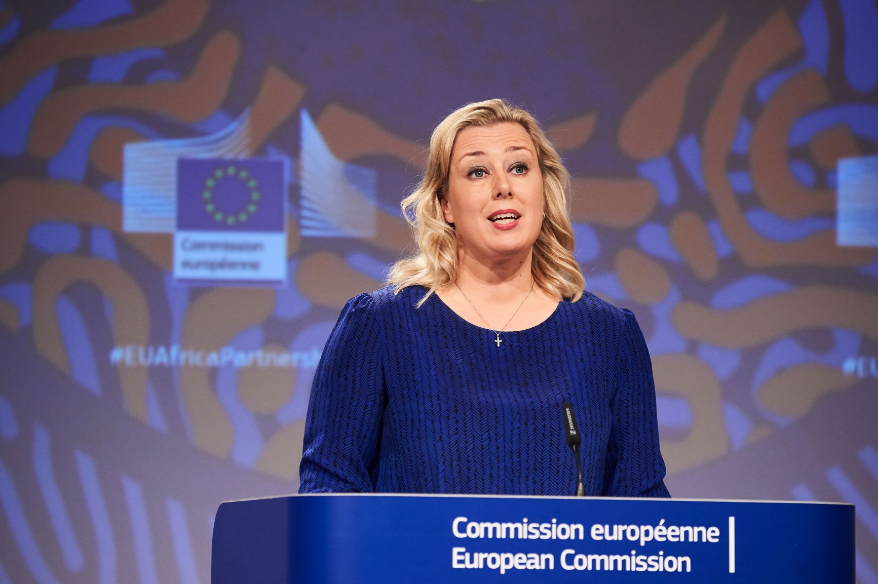 Vaaleahiuksisella naisella on tummansininen asu. Hän seisoo puhujapöntössä, jossa on teksti Comission européenne / European Comission.