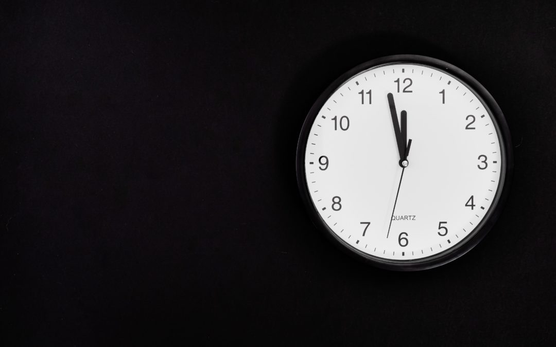 Mustalla seinällä kello, jossa valkoinen tausta ja mustat numerot sekä viisarit. Kello on kuvassa 11.58.
