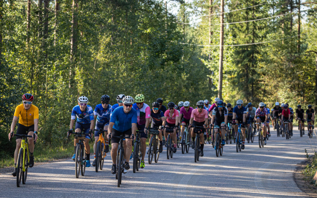 Kymmeniä gravel-pyöräilijöita ajaa metsäisellä maantiellä. Kesä, aurinko paistaa.