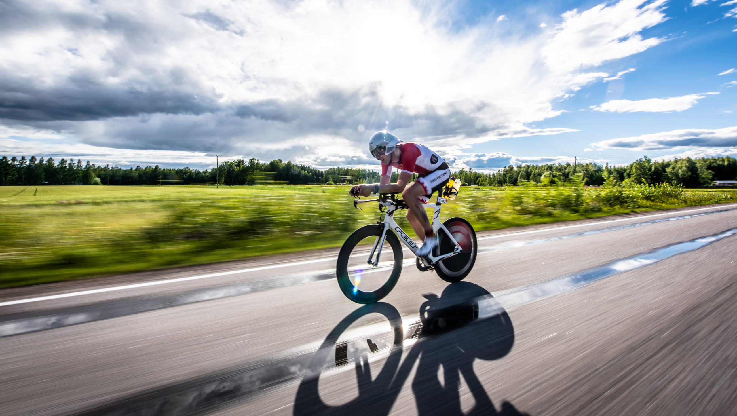 Ironman kilpailu kesällä. Yksi kilpailija ajaa kilpapyörällä maantiellä. Aurinko paistaa pilvien läpi. Taustalla viheralue ja lehtimetsä.
