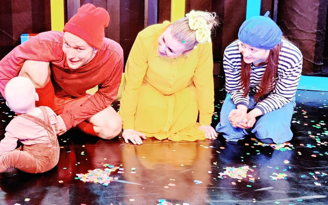 Teatterin lavalla kolme näyttelijää värikkäissä asuissa polvillaan. Heidän edessään istuu taaperoikäinen lapsi.lapsi