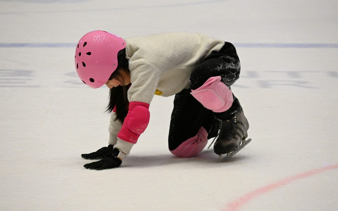 Lapsi luistimet jalassa polvillaan jäällä. Vaaleanpunainen kypärä, kyynär- ja polvisuojat. Valkoinen paita ja mustat housut
