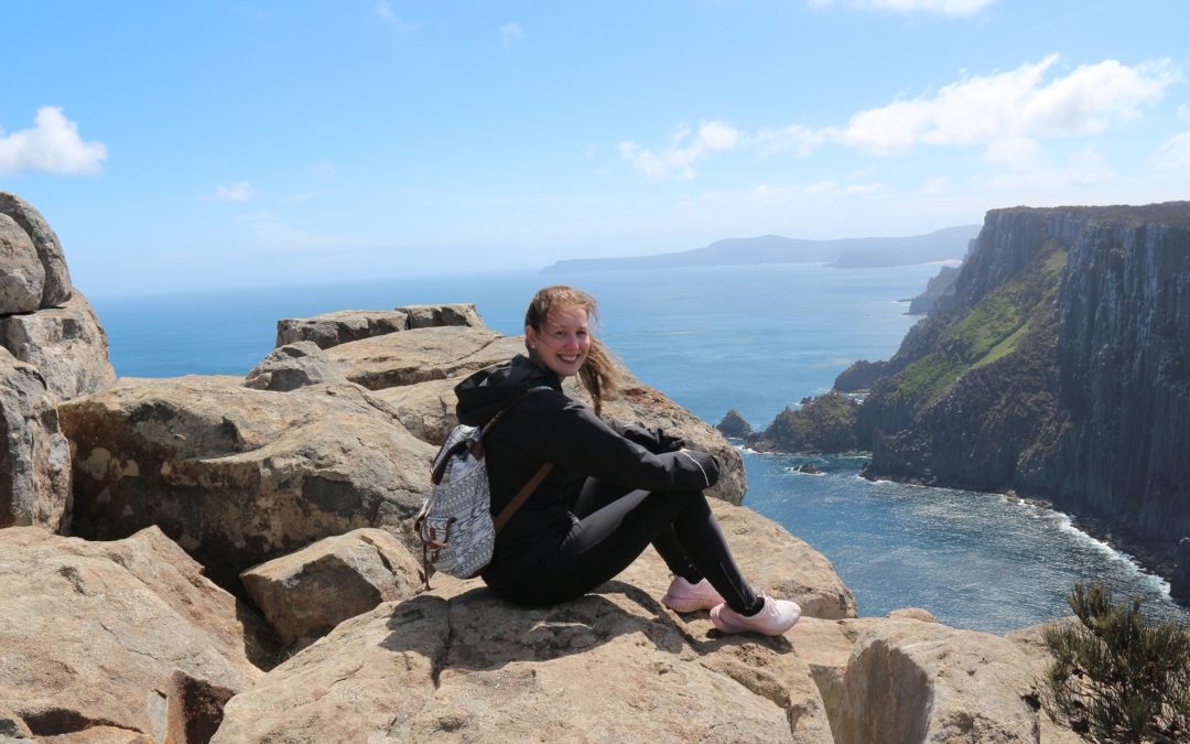 Hymyilevä nainen istuu kalliolla mustissa vaatteissa ja reppu selässä. Taustalla meri.