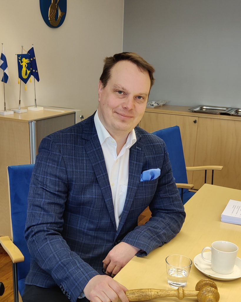 Maakuntahallituksen puheenjohtaja Pekka Komu istuu kokoushuoneessa pöydän ääressä. Pöydällä paperipino, nuija ja kahvikuppi. Taustalla Suomen ja EU-pöytäliput ja Päijät-Hämeen vaakuna.