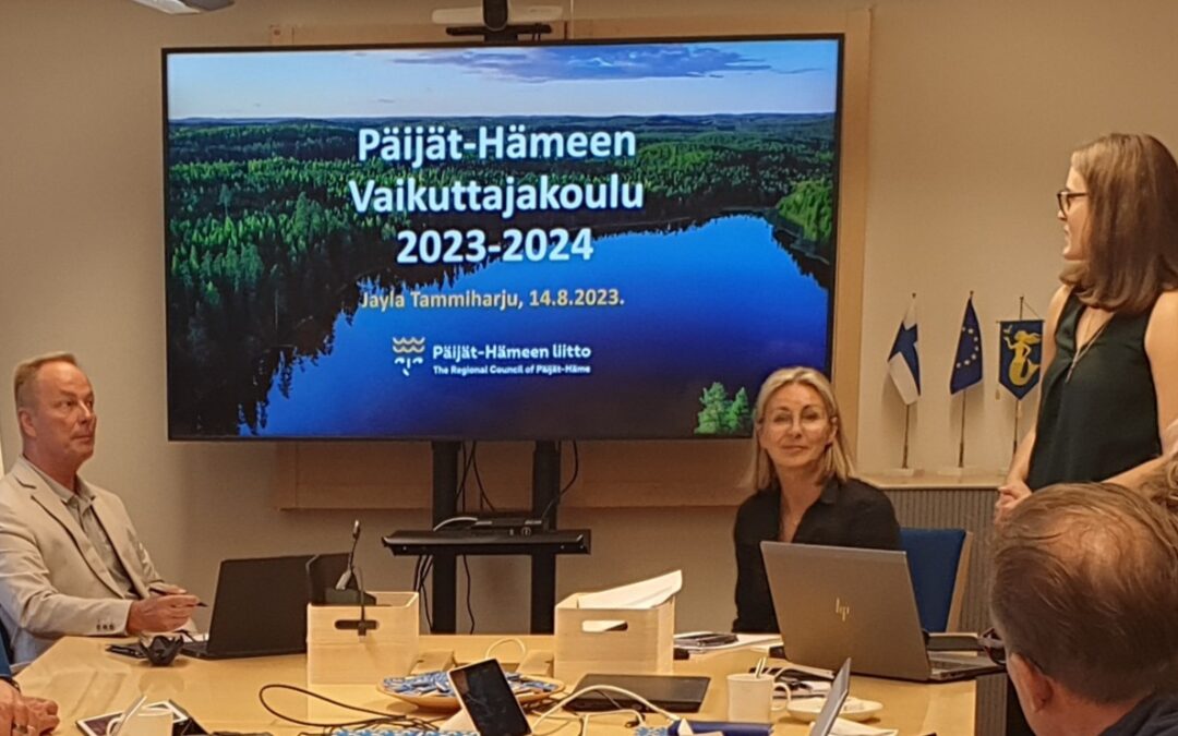 Kokoustila, isolla näytöllä teksti Päijät-Hämeen vaikuttajakoulu 2023-2024. Mies ja nainen uistuvat pöäydän ääressä, nainen pitää seisaaltaan esitystä.