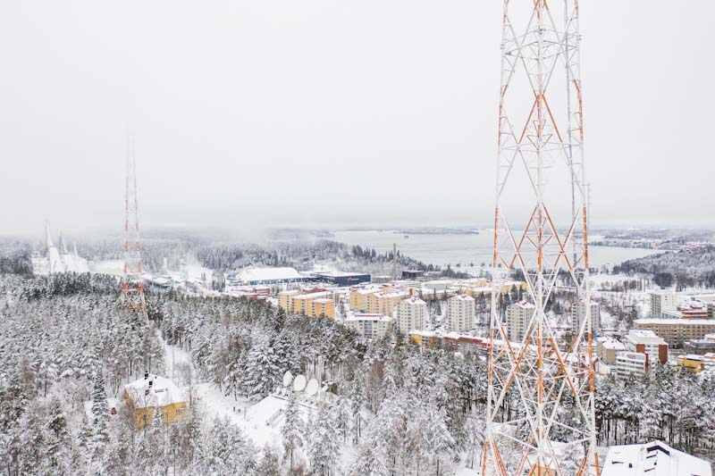 Talvimaisema Lahden kaupungista. Etualalla korkea radiomasto ja taaempana toinen. Mastojen alapuolella kerrostaloja ja järvi. Vasemmassa reunassa kolme hyppyrimäkeä.