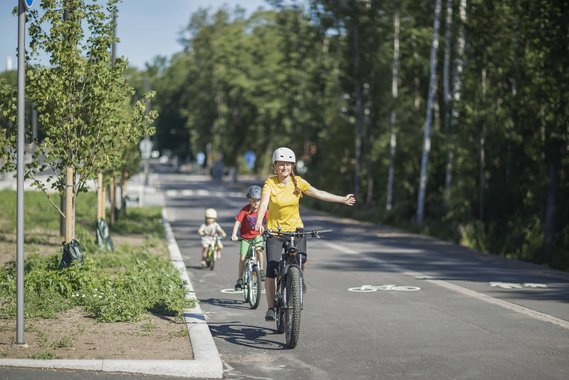Kevyenliikenteen väylällä kesällä kolme eri-ikäistä pyöräilijää, joilla kaikilla kypärä päässään. Nuorin on pieni lapsi. Väylää reunustaa lehtipuut.