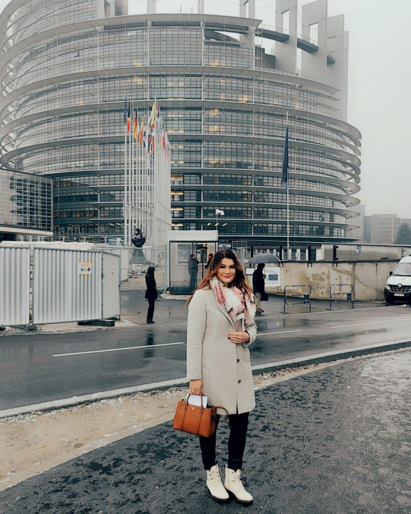 Naiinen seisoo EU-parlamenttirakennuksen edessä Strasburgissa sumuisessa säässä. Iso pyöreä rakennus täynnä ikkunoita. Rakennuksen edessä EU-maiden liput salossa.