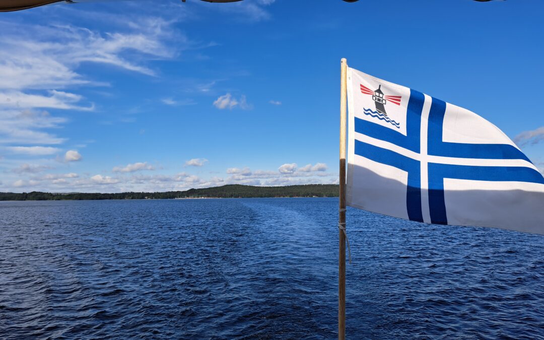 Aurikoinen kesäpäivä järvellä. Veneen perässä lippu, valkoinen pohja, sinisellä rajattu valkoinen risti. Yläkulmassa tyylitelty majakka.